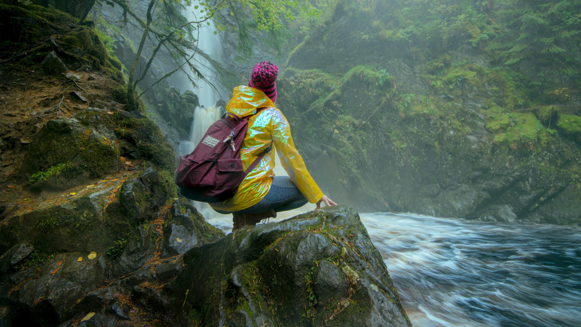 A girl in a yellow jacket sits on a rock at the base of Plodda Falls